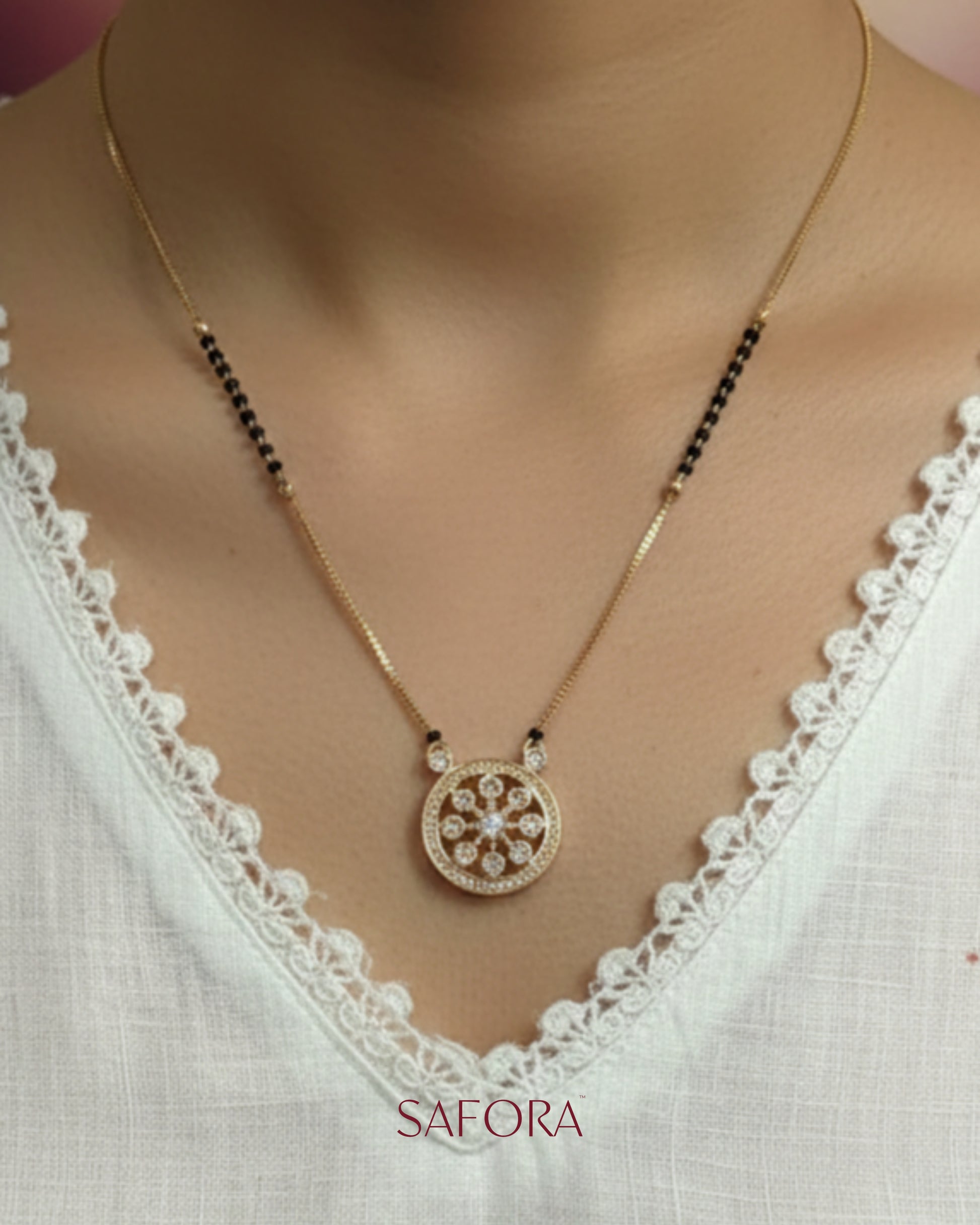 Gold necklace with a decorative pendant on a white lace top, branded 'Safara'.