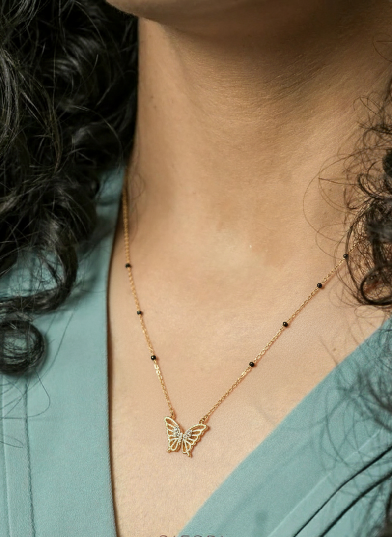 Gold butterfly necklace worn by a person with a teal garment, brand 'SAFORA' visible.